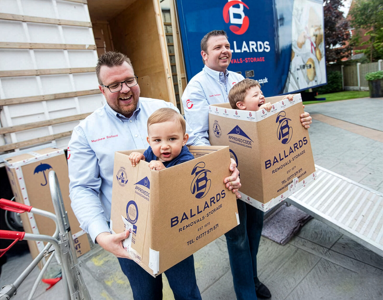 Ballards directors Matt and Joe carrying their young children in boxes in front of a removals truck - family business - Ballards