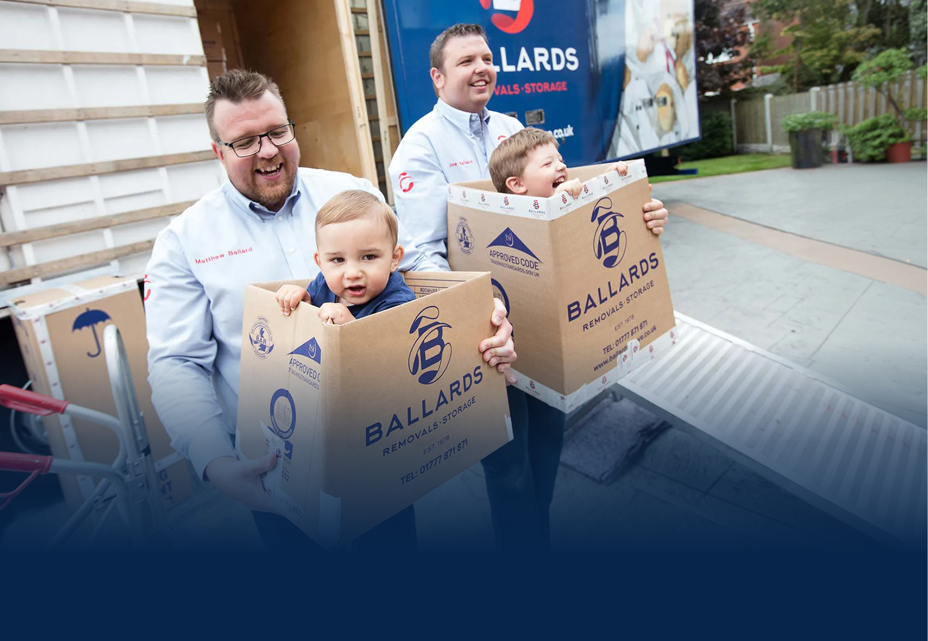 Ballards directors Matt and Joe carrying their young children in boxes in front of a removals truck - family business - Ballards