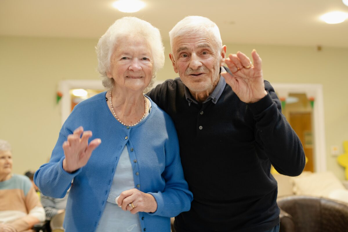 Two older people wave to the camera smiling