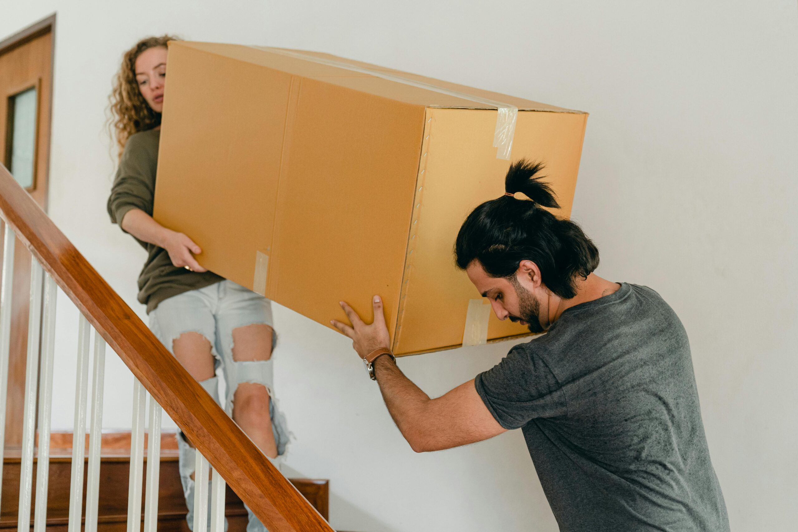 a man and woman carring either end of a large box down the stairs of their home