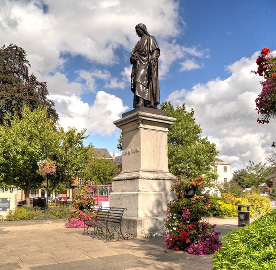 A stone statue of Sir Isaac Newton on St Peter’s Hill in Grantham, showing the figure standing on a plinth with detailed sculpting and surrounding greenery visible in the background.