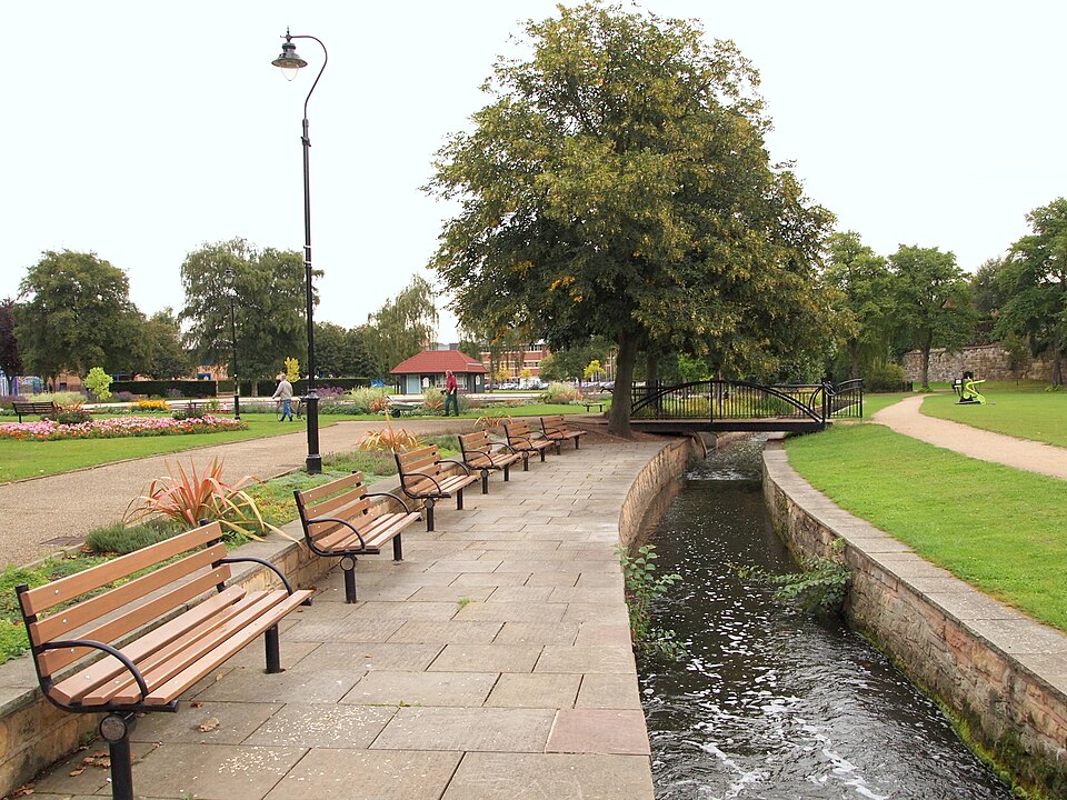 Benches in Titchfield Park, Mansfield facing a small stream, with trees and greenery creating a calm, scenic park setting.