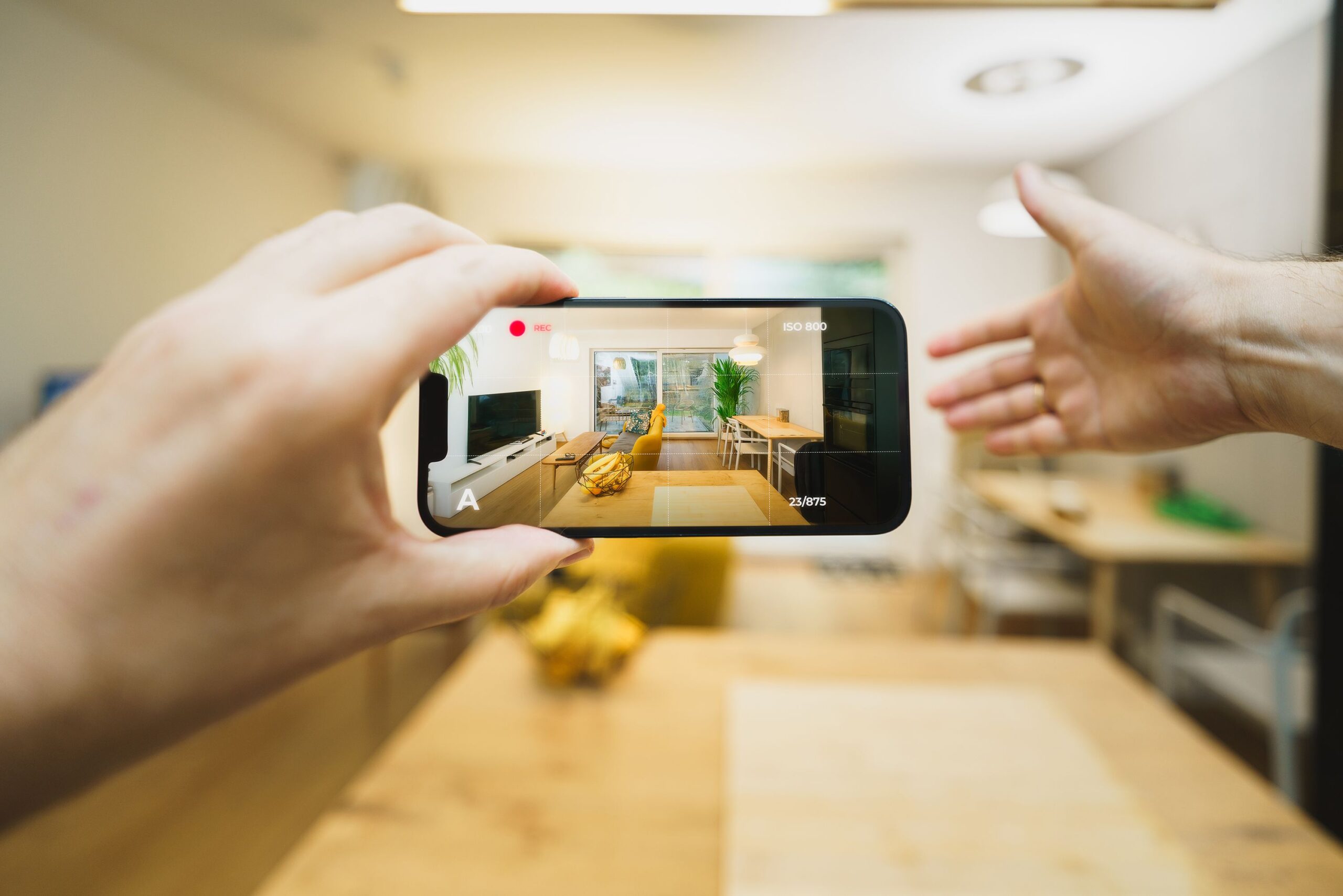 Hands holding a smartphone screen, showing a recording of a well‑lit living room with furniture and a large window.