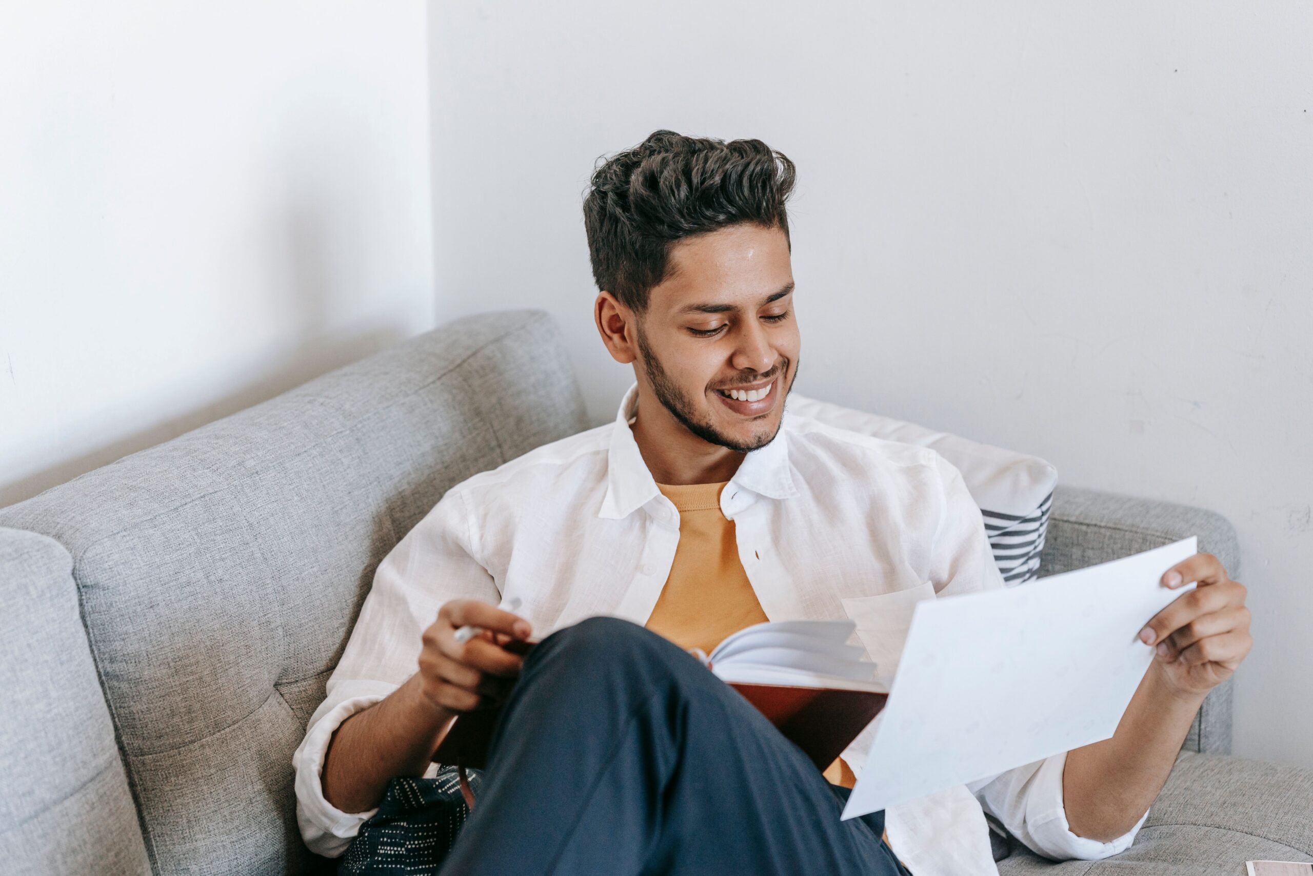 Man smiling as he looks at a printed sheet of paper while sitting on a sofa with a book on his lap.