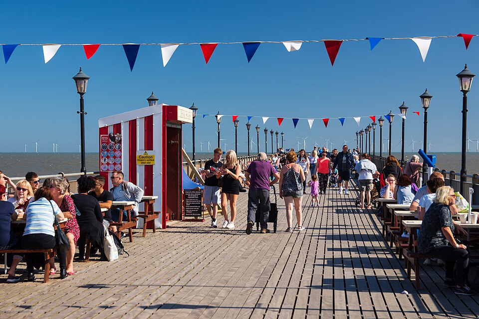 People walking and relaxing on Skegness Pier on a sunny day, with colourful bunting overhead, picnic tables along the wooden deck and wind turbines visible on the horizon.