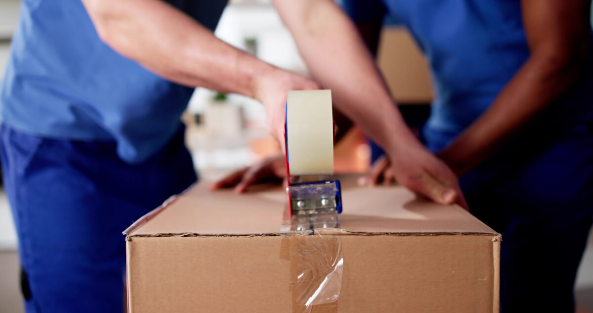 Two people in blue clothing seal a large cardboard box with clear packing tape, using a tape dispenser while preparing items for transport or storage.